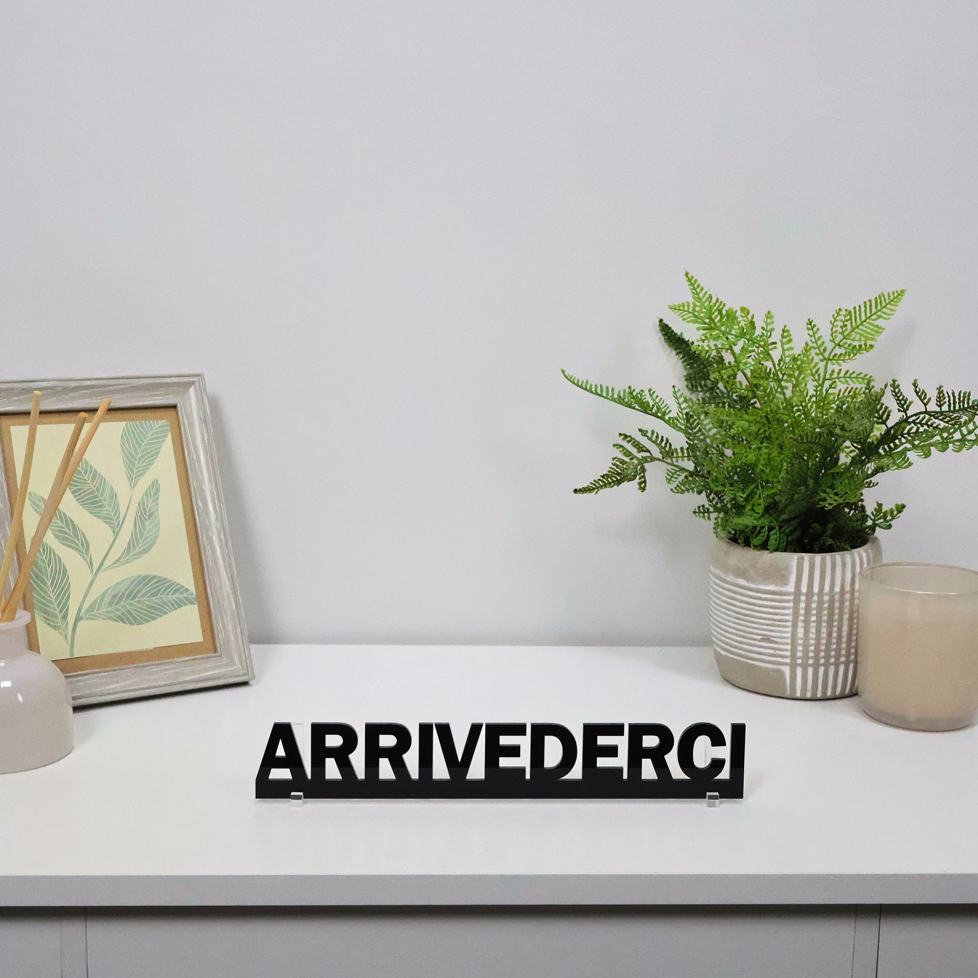 Black ARRIVEDERCI sign displayed freestanding on a desk beside a plant.