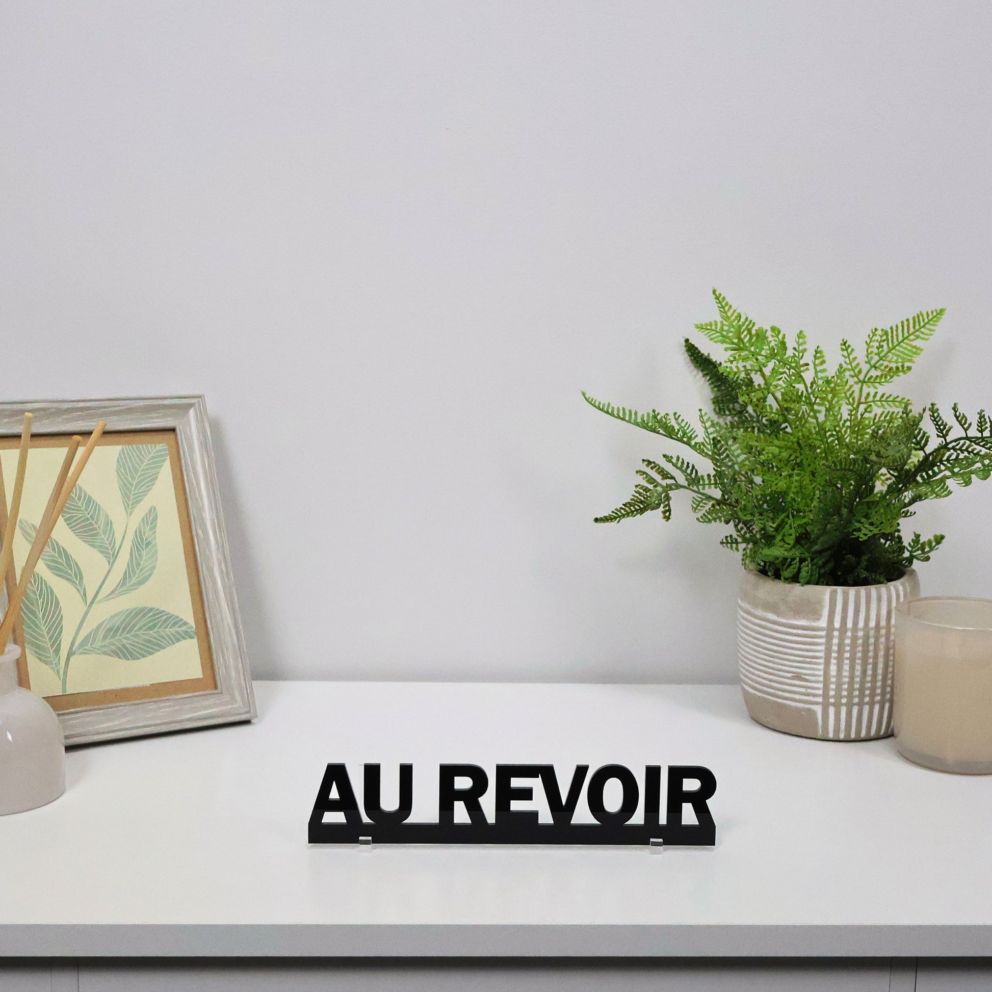 AU REVOIR Door Topper freestanding on a console shelf beside candles and books.