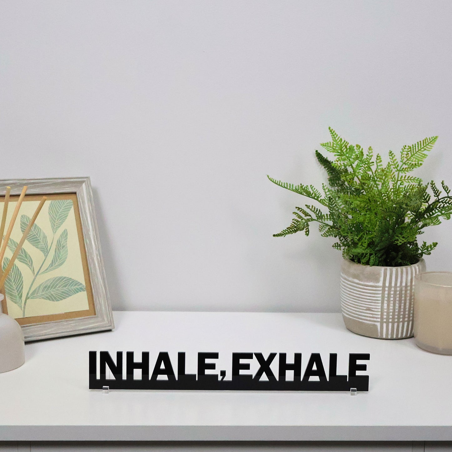Calming acrylic sign freestanding on a shelf beside a plant and candle