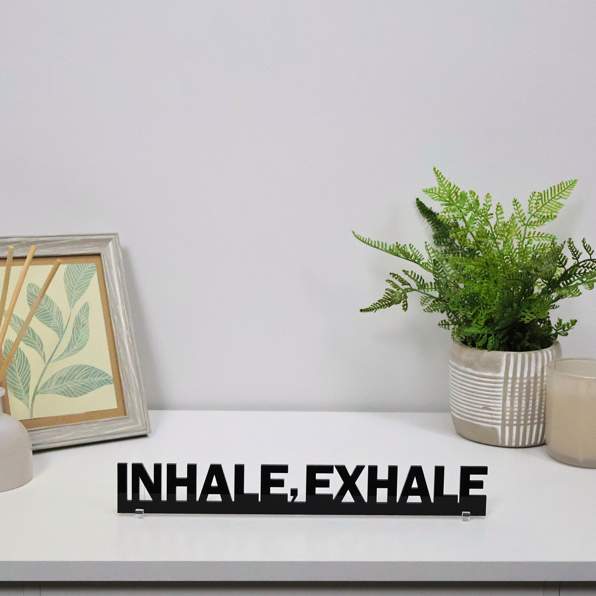 Calming acrylic sign freestanding on a shelf beside a plant and candle